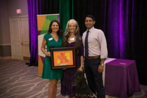 Three people smile at the camera while recieving an award. Service Dog, Frank, is also featured.