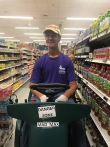 Man sits behind wheel of floor waxing machine inside a grocery store.