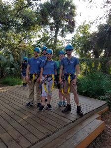 Five people stand on a low platform with harnesses around thier waists.